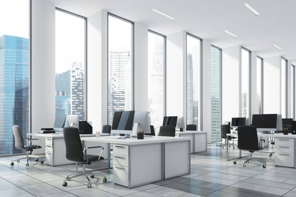 A modern open-plan office with white desks, black chairs, computers, and large windows overlooking a city.