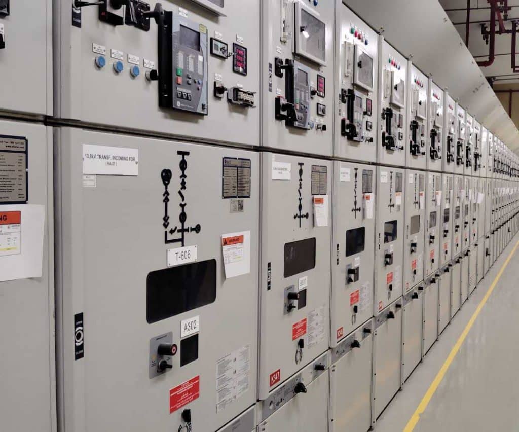 A long row of gray electrical panels with various meters, switches, and labels, including "13.8KV TRANSF. INCOMING FOR."