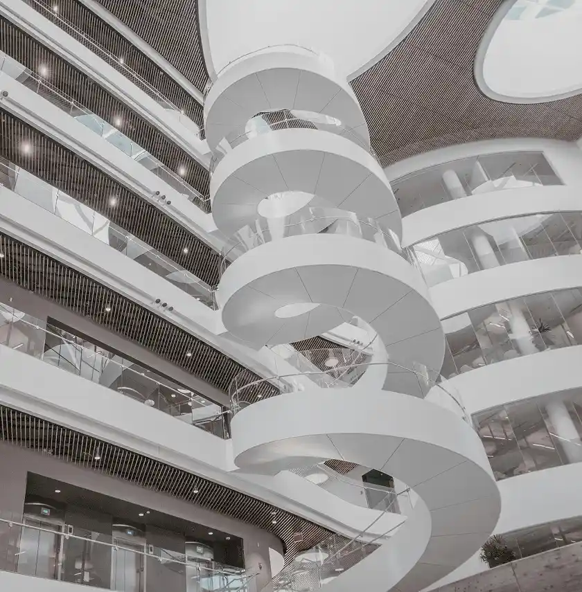 A modern building atrium features a large white spiral staircase, glass railings, and multiple floors with balconies.