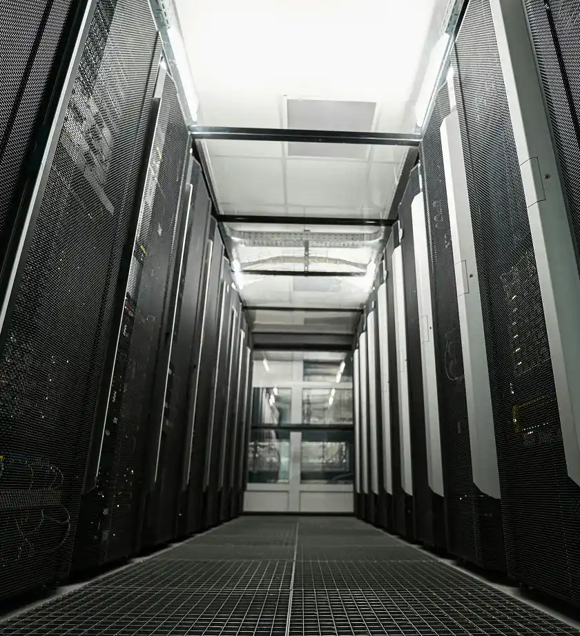 A low-angle view down a narrow aisle between rows of tall, dark server racks with a grated floor.