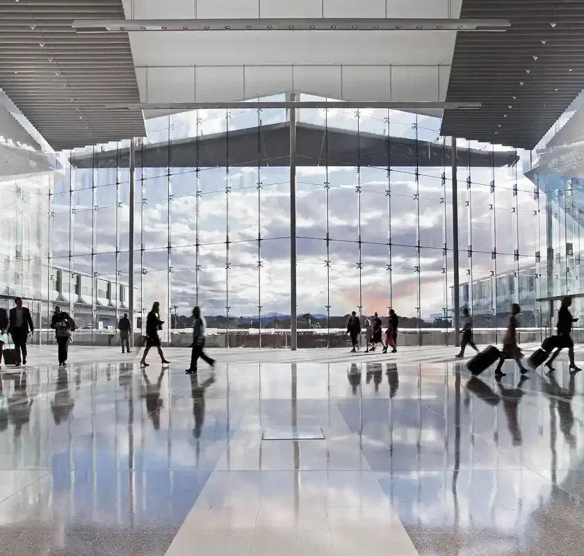 People walk across a reflective floor in a modern building with a large glass wall and cloudy sky outside.